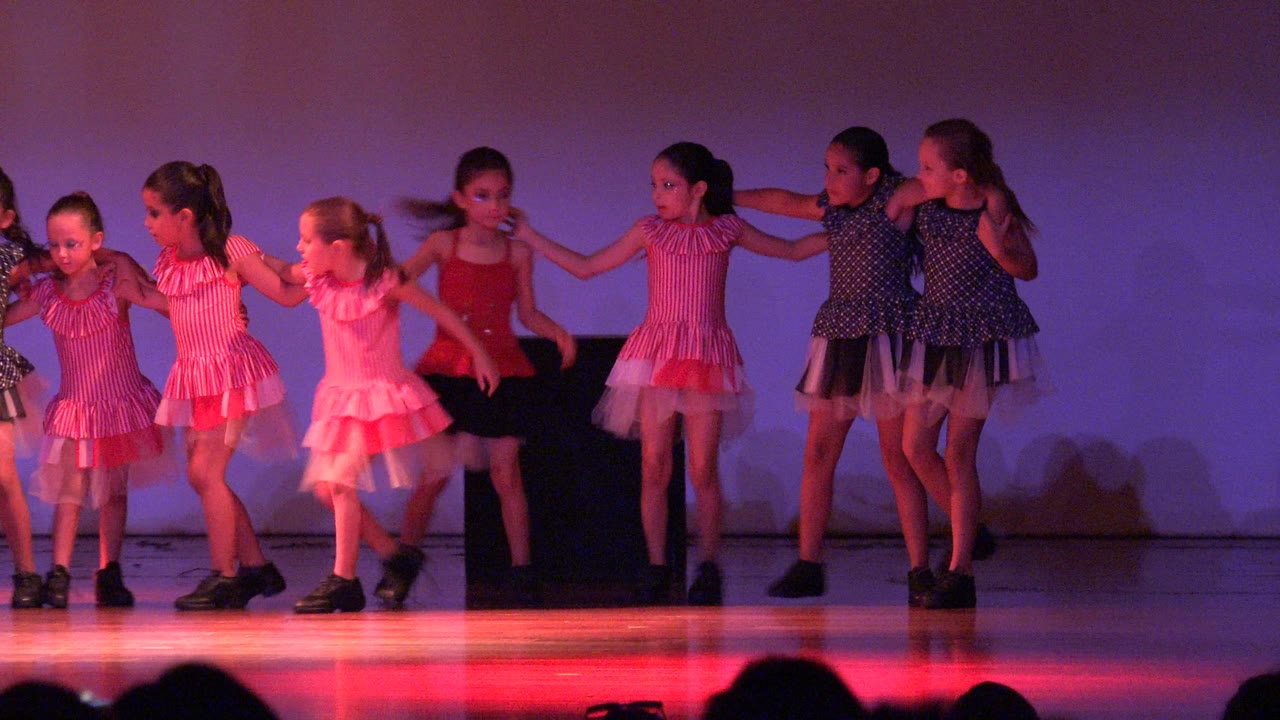 Young girls in pink and dark dance outfits stand across the stage in San Jose del Cabo BCS Mexico, with red light reflected on the floor and audience silhouettes at the bottom.