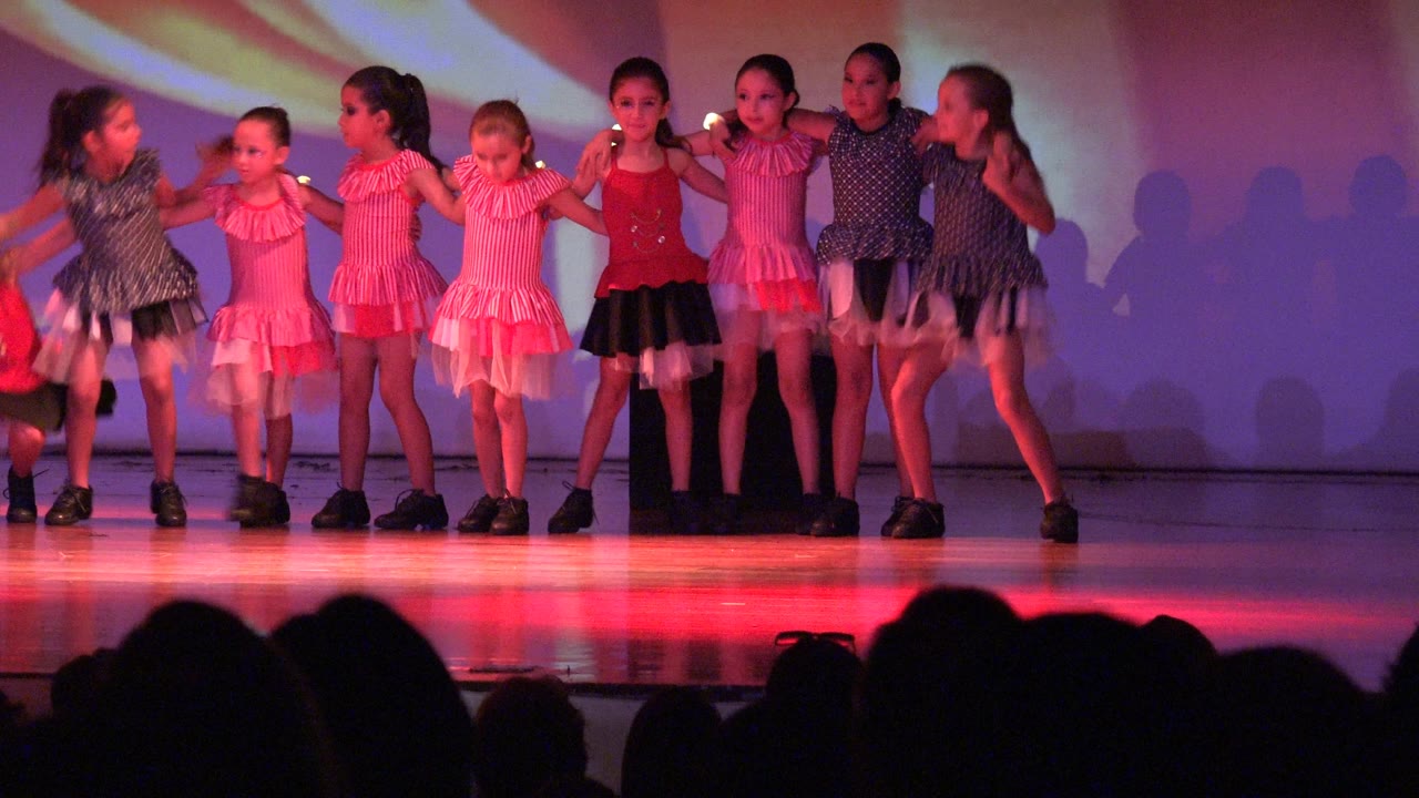 Young girls in San Jose Del Cabo BCS Mexico stand hand in hand on a stage under red and purple lighting, with audience silhouettes at the bottom of the frame.