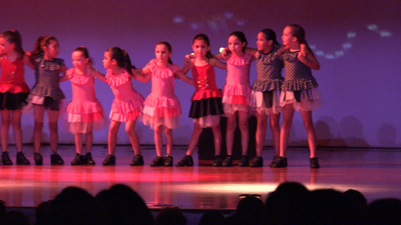 Girls in dresses lined up across a stage under colored light.