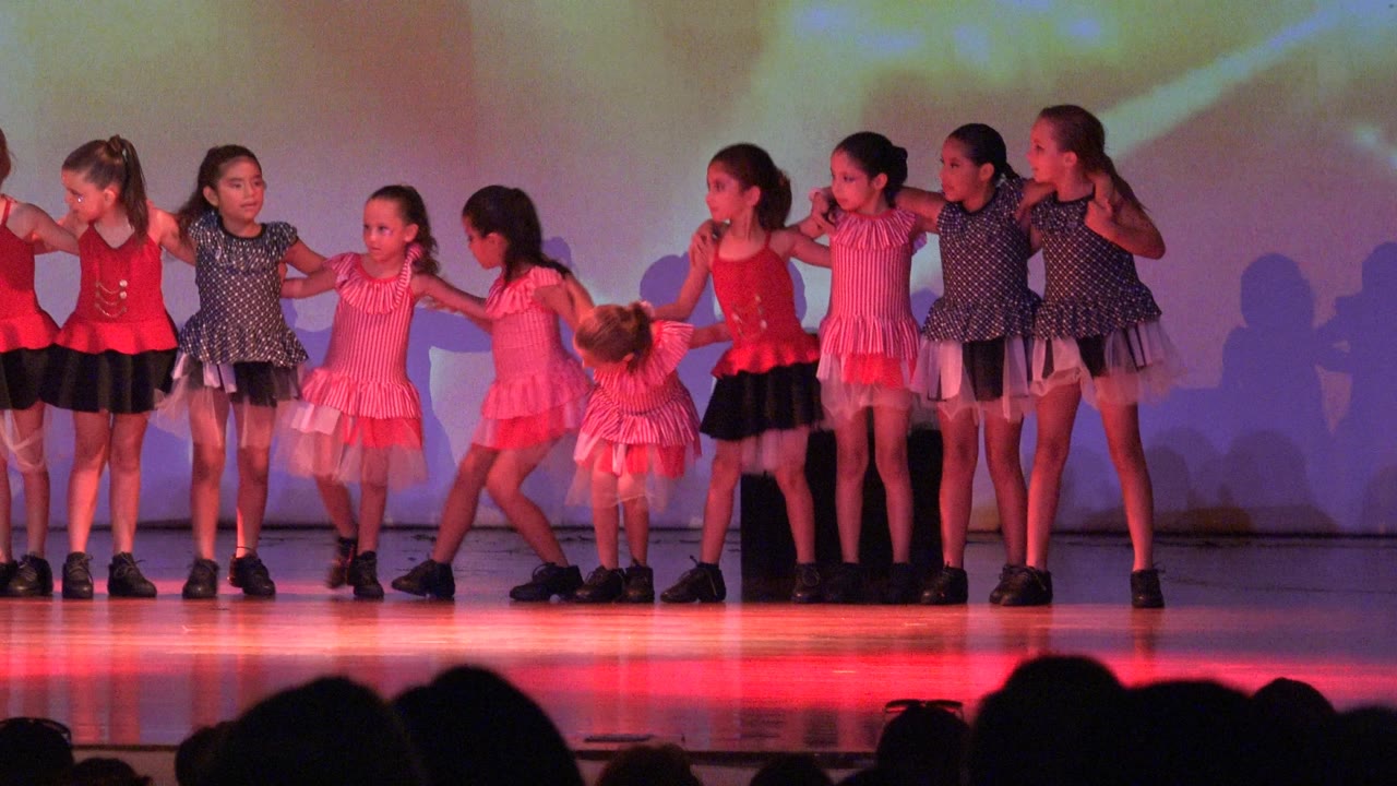 During the San Jose Del Cabo BCS Mexico performance, one girl bends forward near center while the rest of the children keep their handhold line under warm stage light.