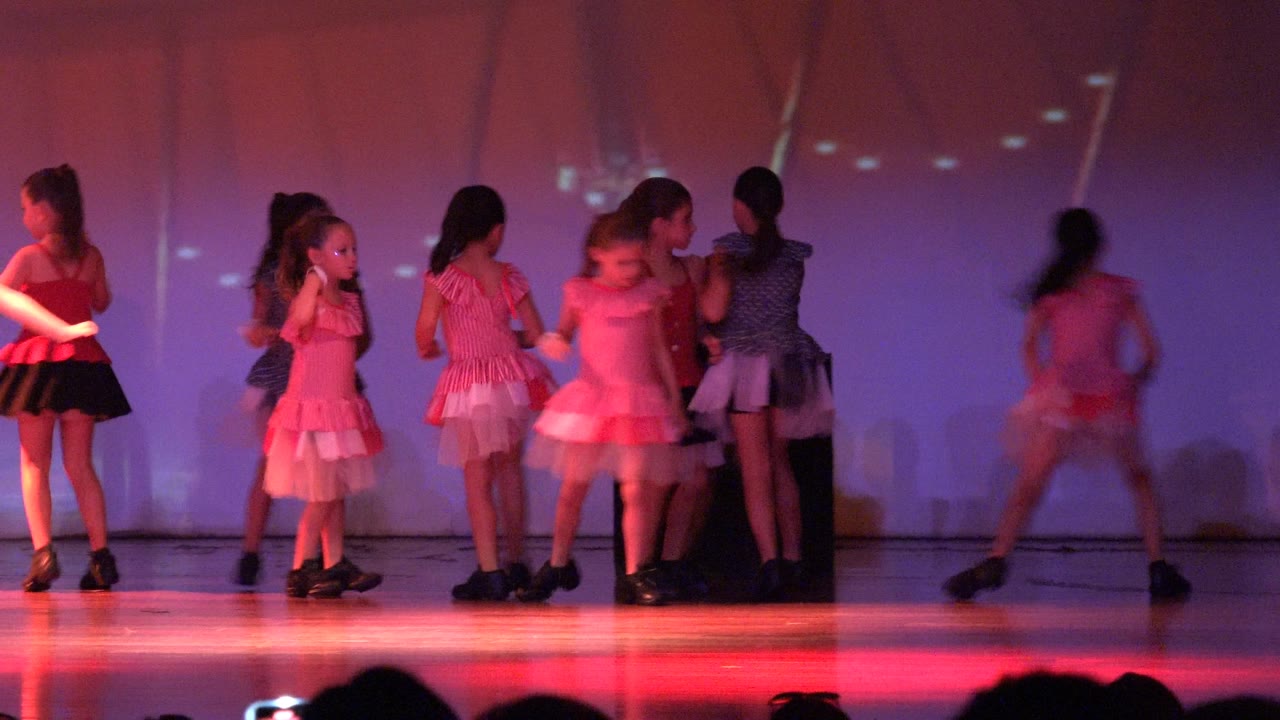Wide stage frame in San Jose del Cabo, Mexico shows young girls in pink and red dance outfits gathering and moving under purple lighting, with audience silhouettes along the bottom edge.