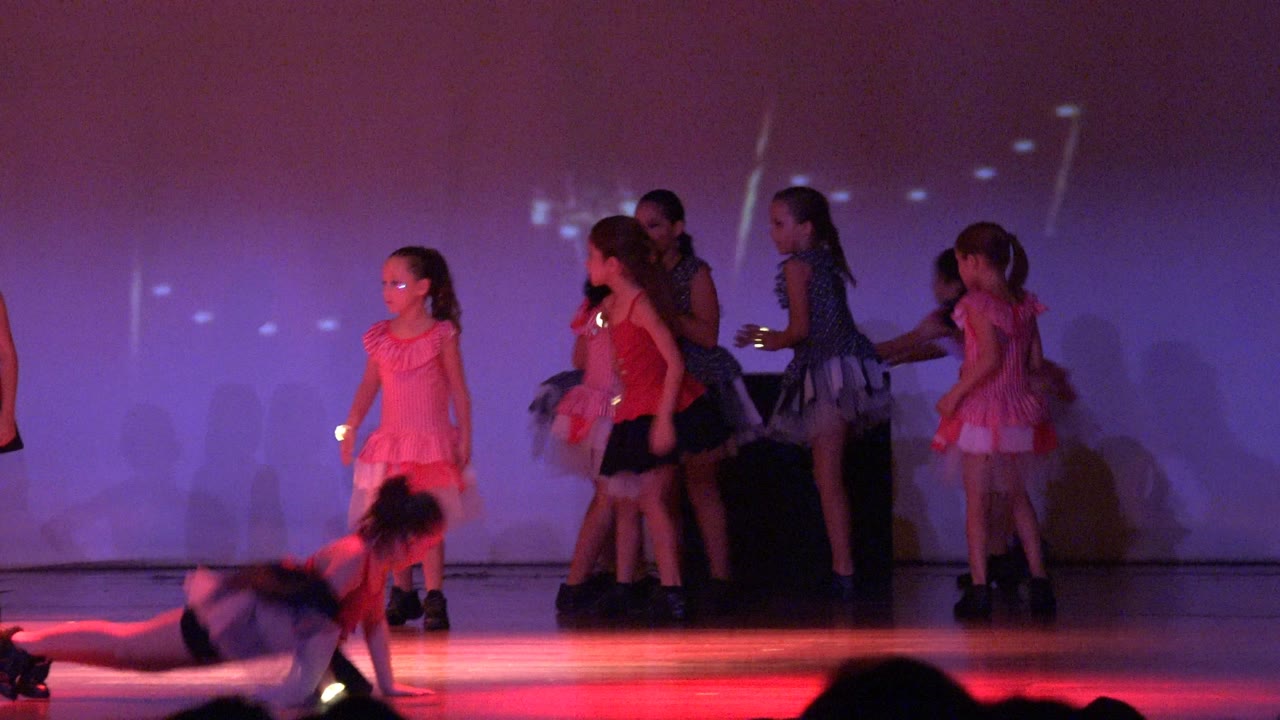 A child lies or slides across the front of the stage in San Jose del Cabo as several girls in pink, red, black, and polka-dot costumes stand and shift under purple light.