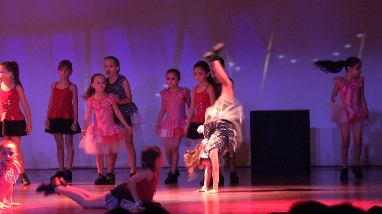 Children in colorful costumes perform on a stage in San Jose del Cabo, Mexico, with one dancer upside down in a cartwheel and others standing or seated under red and purple light.