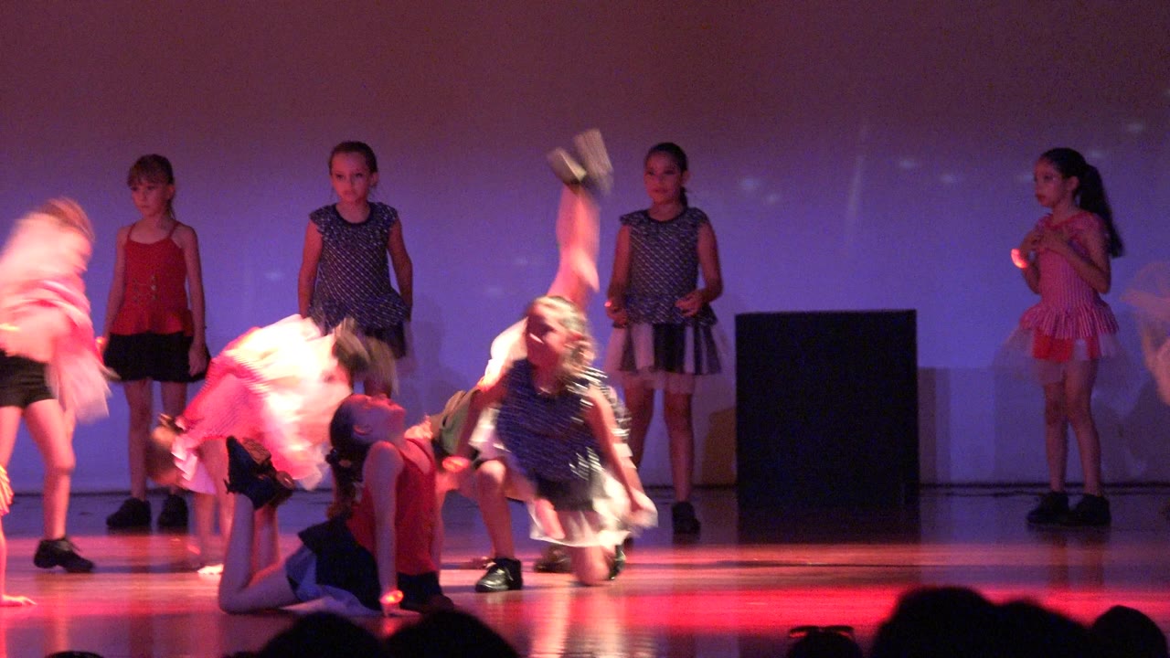 Children in pink, red, navy, and black costumes perform on a red-lit stage in San Jose del Cabo, Mexico, with one dancer blurred mid-move near the center and others watching or holding choreography.