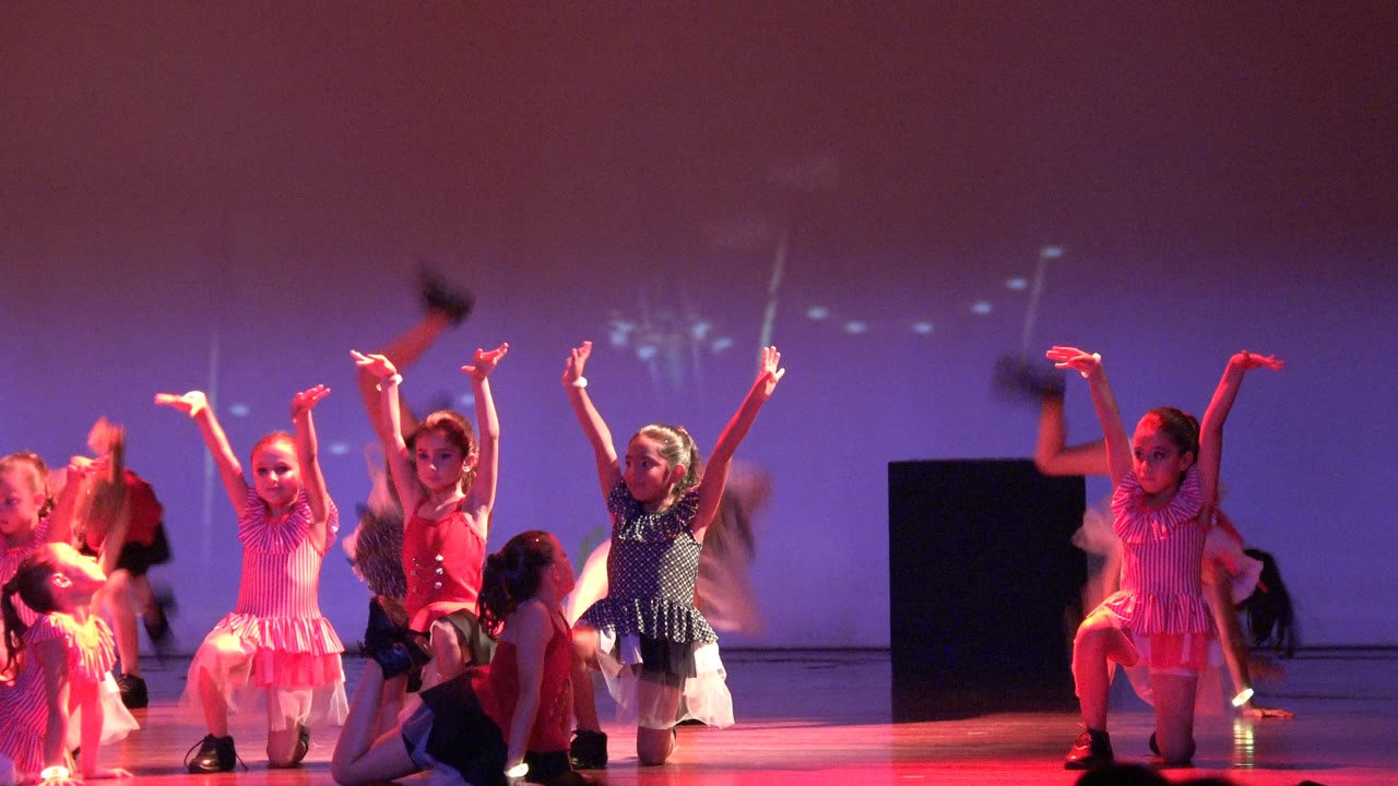 Young girls in colorful costumes perform on a stage in San Jose del Cabo, Mexico, with several arms raised against a purple backdrop.