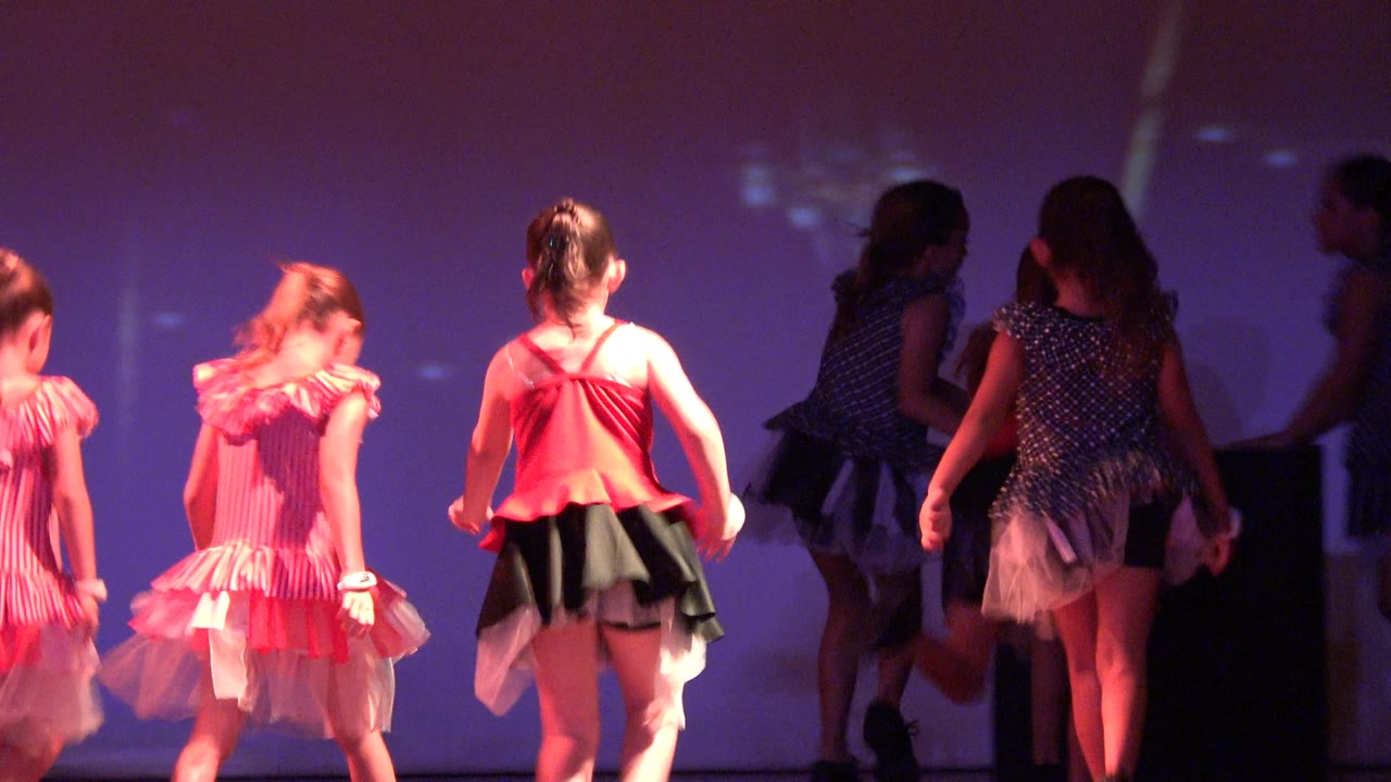 Young girls in pink and blue dance costumes move across a purple-lit stage in San Jose del Cabo BCS Mexico, with several backs turned during the routine.