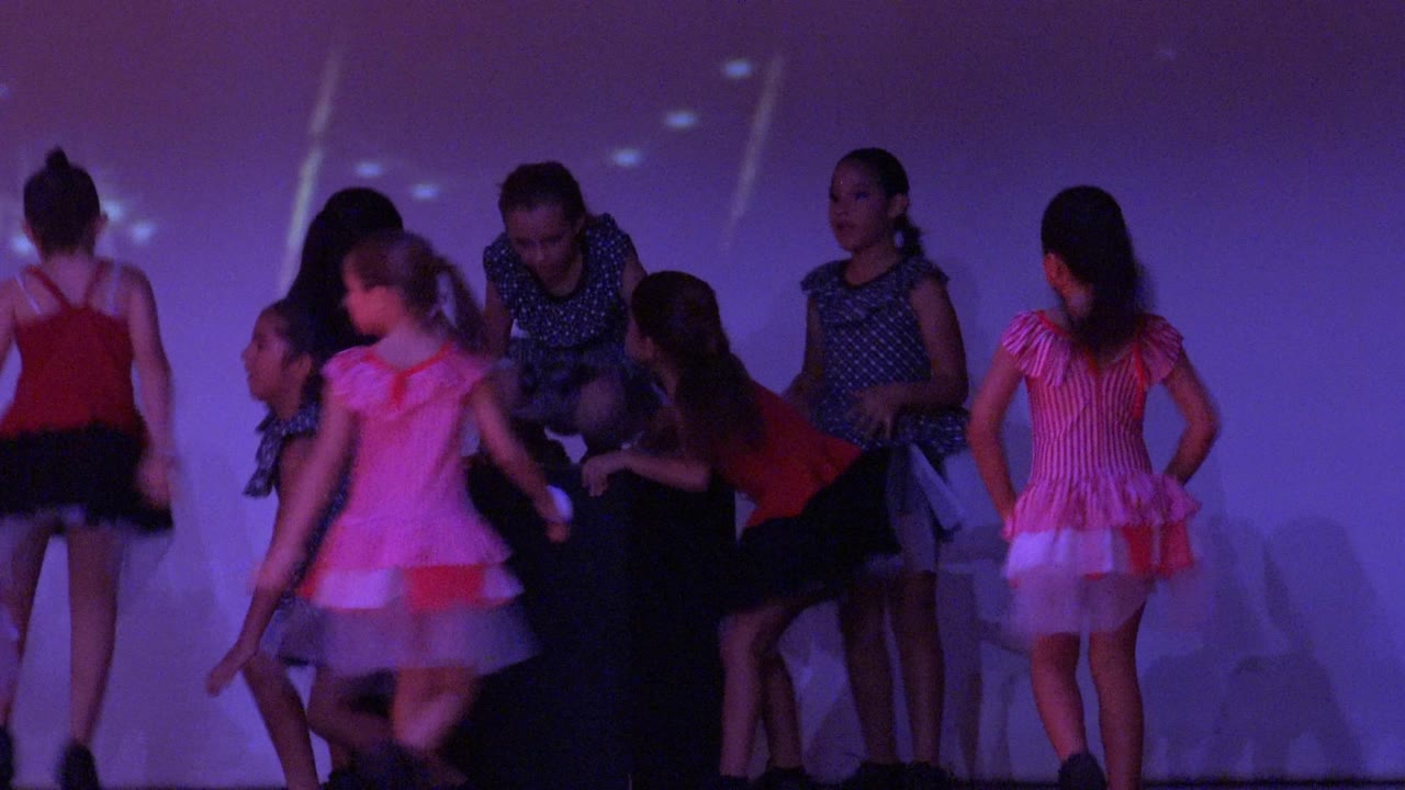 Young dancers continue the stage routine in San Jose del Cabo BCS Mexico, with several girls facing sideways and a bright pink costume standing out against the blue-purple background.
