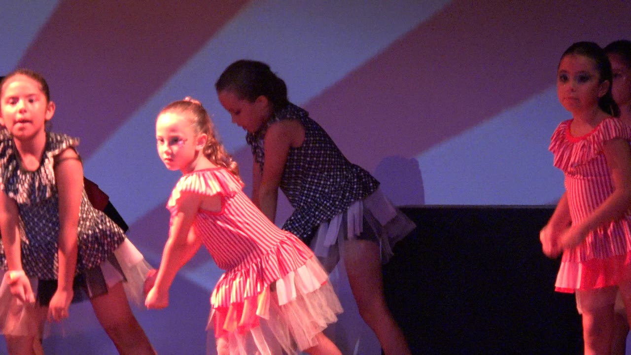 Young girls in pink striped and dark polka-dot costumes perform on a stage in San Jose del Cabo, Mexico, with purple and red lighting across the backdrop.