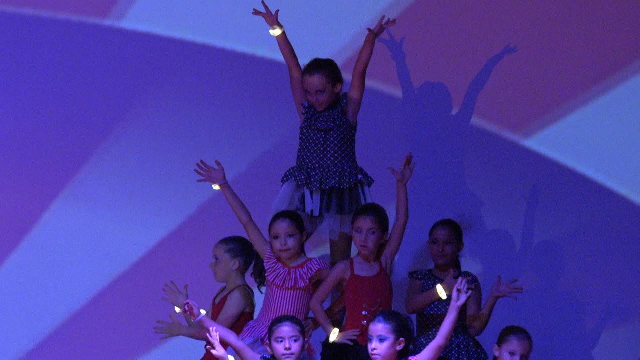A group of children maintains a synchronized pose on the stage in San Jose del Cabo BCS Mexico, with the elevated dancer centered near the top and colored lights on wrists.