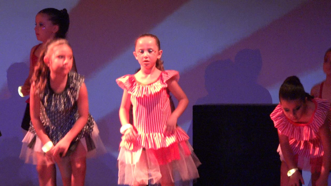 Young girls in red, pink, black, and white dance costumes stand across a stage in San Jose del Cabo, Mexico, with a purple and white backdrop behind them.