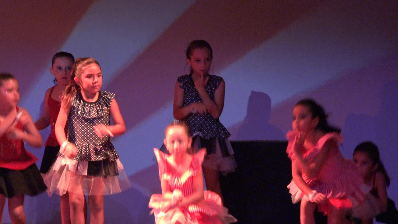 Several young girls in pink and dark dresses stand spaced across the stage in San Jose del Cabo, Mexico, with striped backdrop light and a dark block behind them.