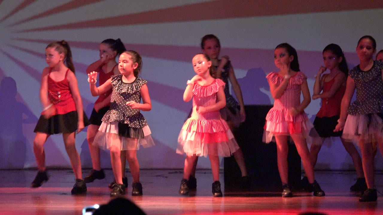 Young girls perform a dance routine on a San Jose Del Cabo BCS Mexico stage, with red and pink costumes, black shoes, and a striped backdrop behind them.