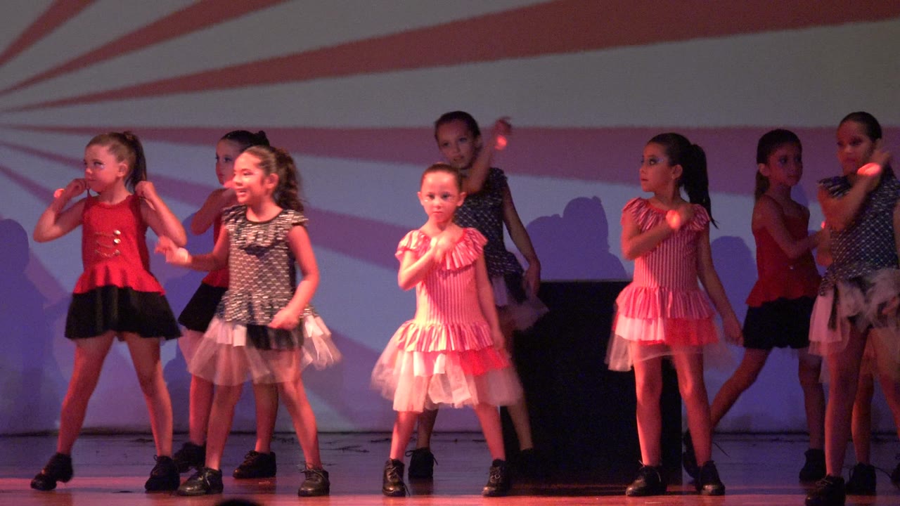 Girls continue the stage routine in San Jose Del Cabo BCS Mexico, with one child in a pink ruffled dress near the front and others grouped behind under red-purple lighting.