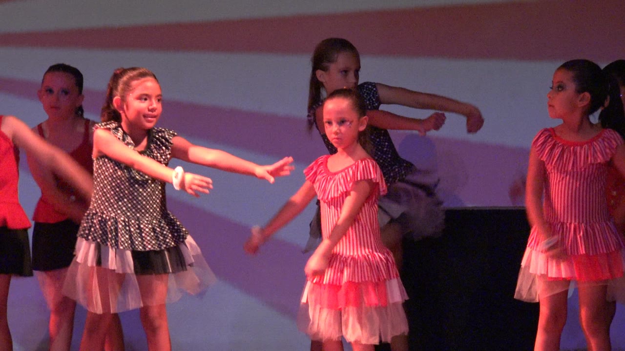 Young girls perform on a stage in San Jose Del Cabo BCS Mexico, with a child in a dark polka dot dress reaching forward beside dancers in pink dresses.