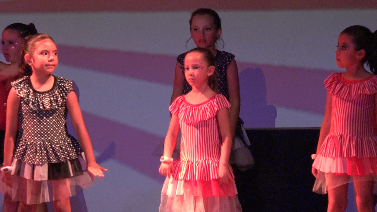 A girl in a dark polka dot dress stands near the center while other young dancers in pink dresses move around her on a lit stage in San Jose Del Cabo BCS Mexico.