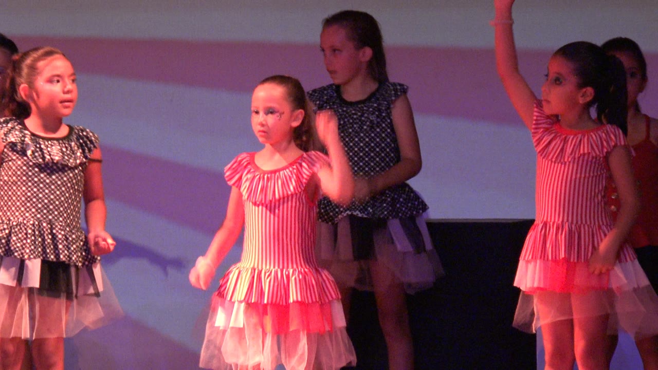 Young girls in San Jose del Cabo BCS Mexico stand on a stage in pink striped and dark polka dot dresses as the dance routine begins under red-pink lighting.