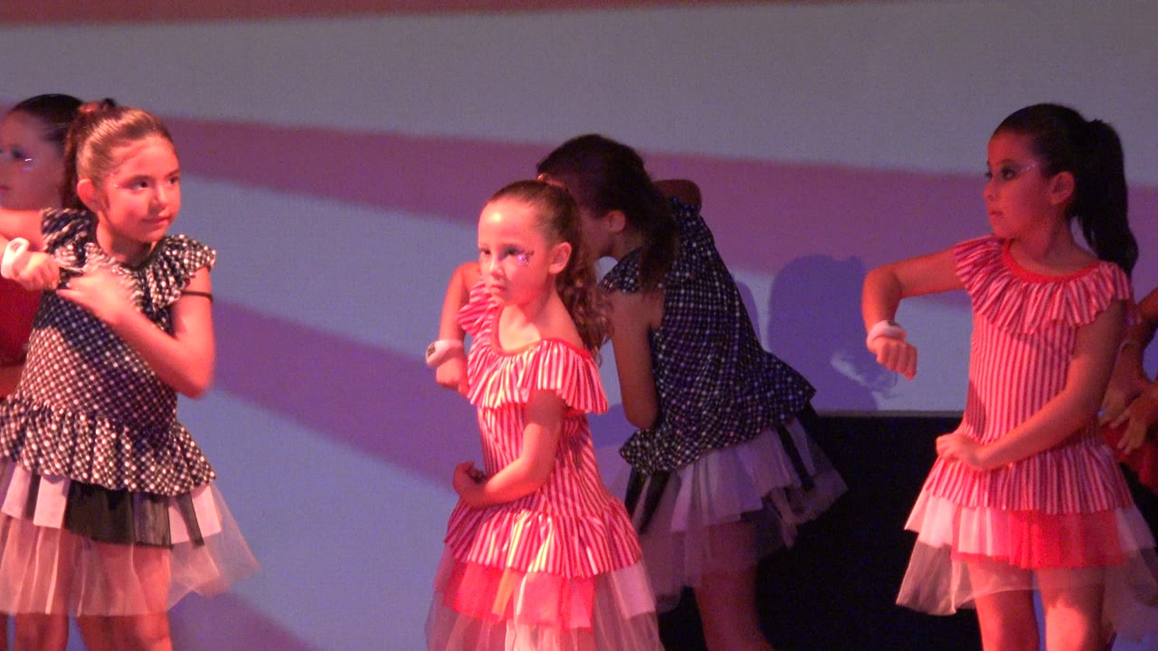 A young girl in a pink striped dress stands near center in San Jose del Cabo BCS Mexico while other children in polka dot tulle dresses continue the stage routine around her.