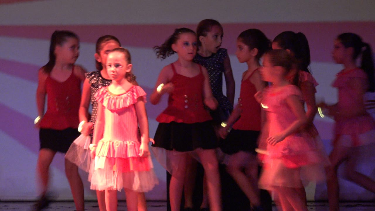 Young girls in red and pink dance costumes stand and move on a stage in San Jose Del Cabo BCS Mexico, with pink light and a striped backdrop behind them.