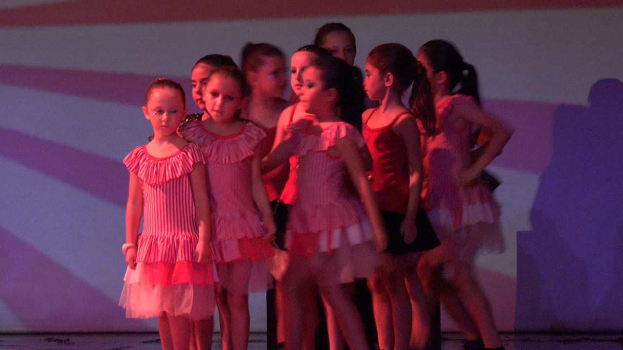 Girls in dance costumes grouped at center stage under pink light.