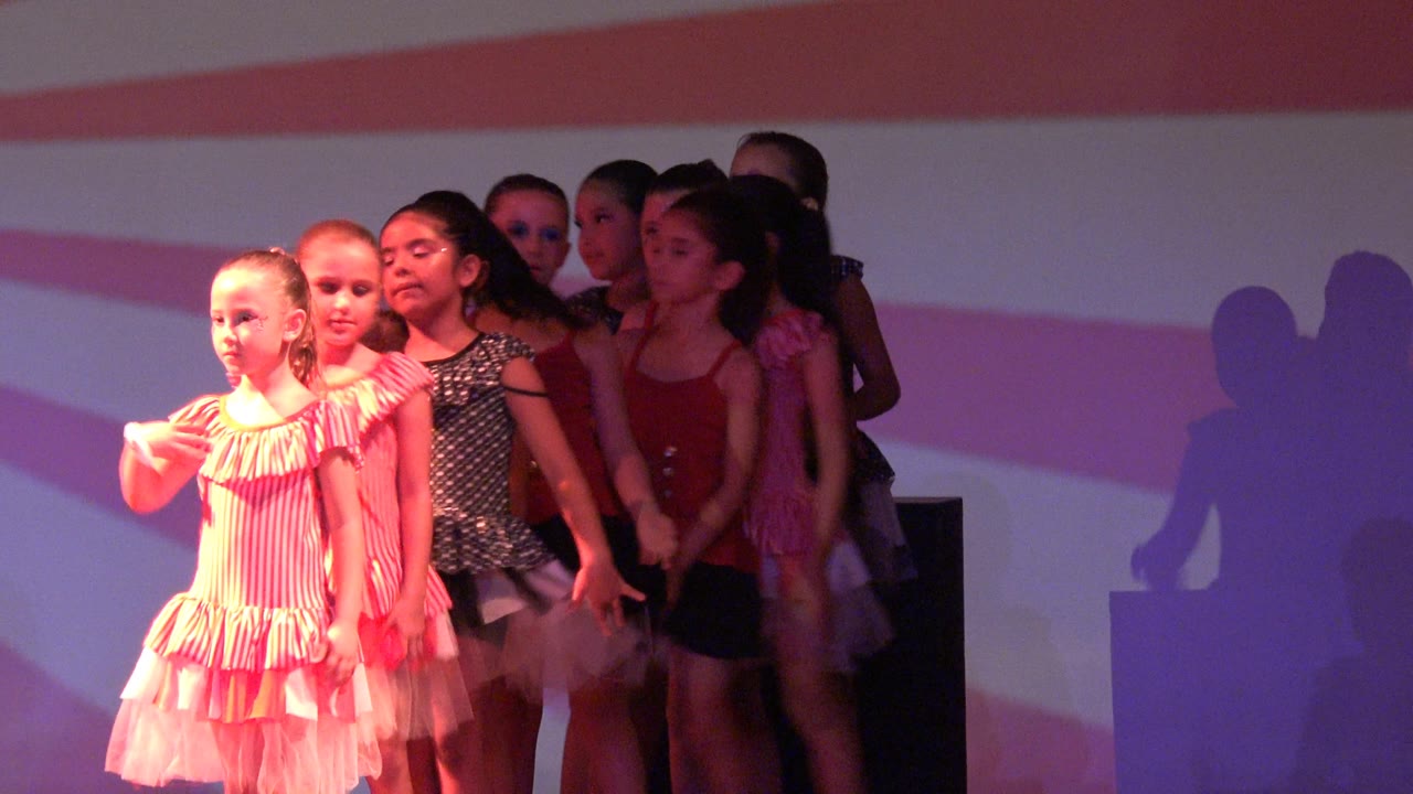 Young girls in matching pink dresses and darker costumes stand in a stage line in San Jose del Cabo BCS Mexico, with red and white stripes projected behind them.