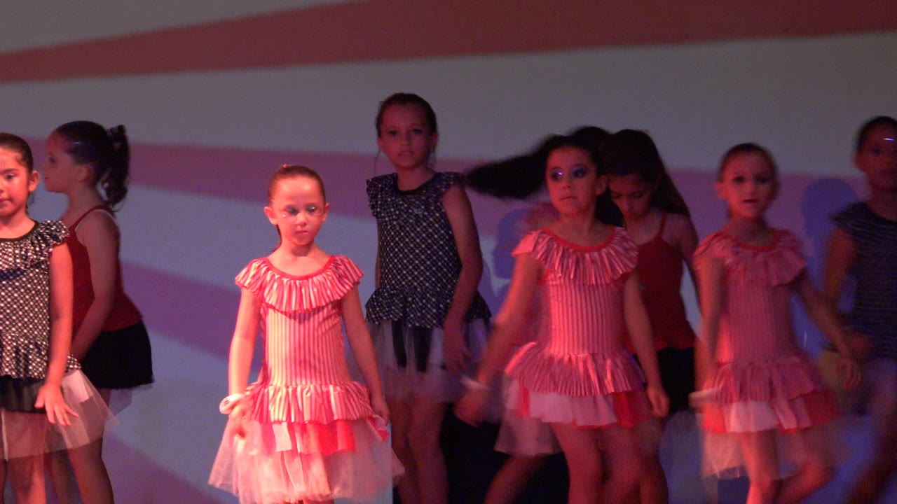 Young girls in San Jose Del Cabo BCS Mexico stand in formation on a stage, with pink striped dresses, dark polka dot costumes, and a pink and white striped backdrop.