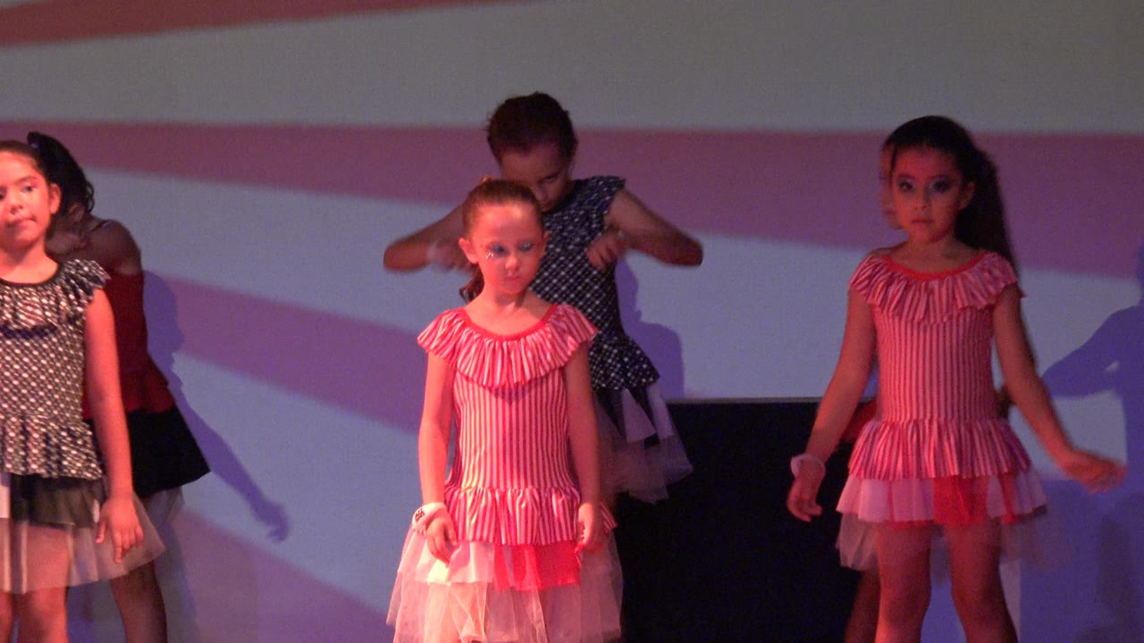 Two young girls in San Jose Del Cabo BCS Mexico stand near the front of the stage, with other dancers behind them and a pink and white striped background across the rear wall.