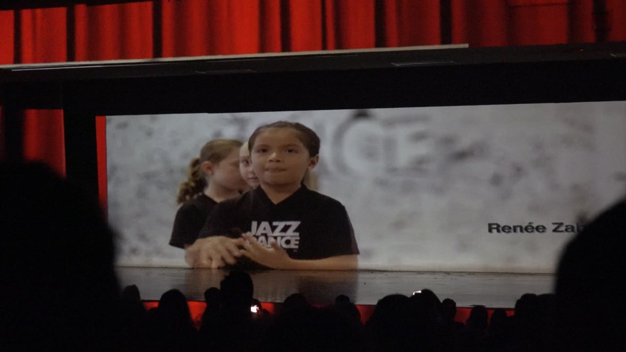 At 0:00, children in black Jazz Dance shirts are displayed on a large theater screen in Mexico, with red curtains behind and dark audience silhouettes below.