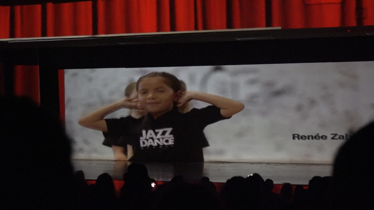 Child dancer raises both hands near her head on the stage screen.