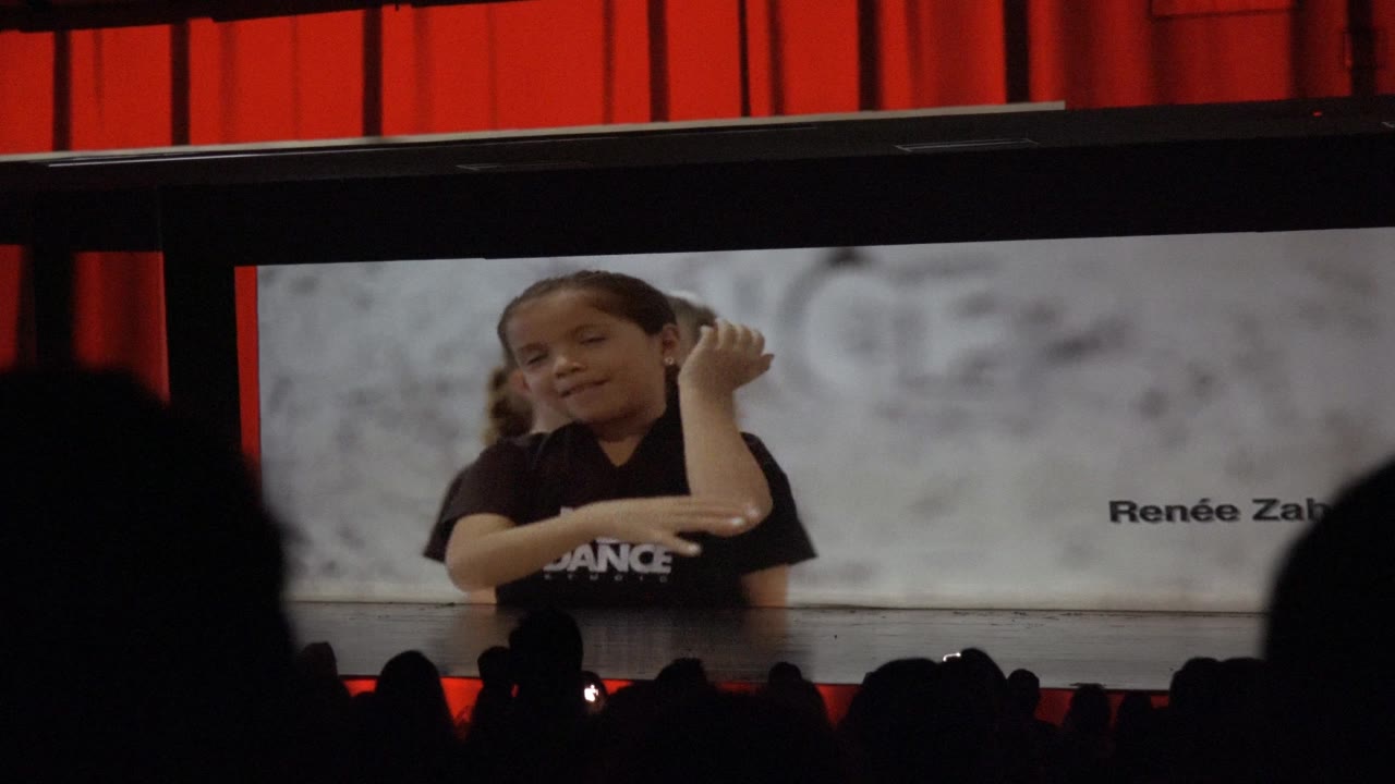 At 0:02, a child dancer on the theater screen in Mexico gestures with one hand, with another child partly behind her and red curtains framing the stage.