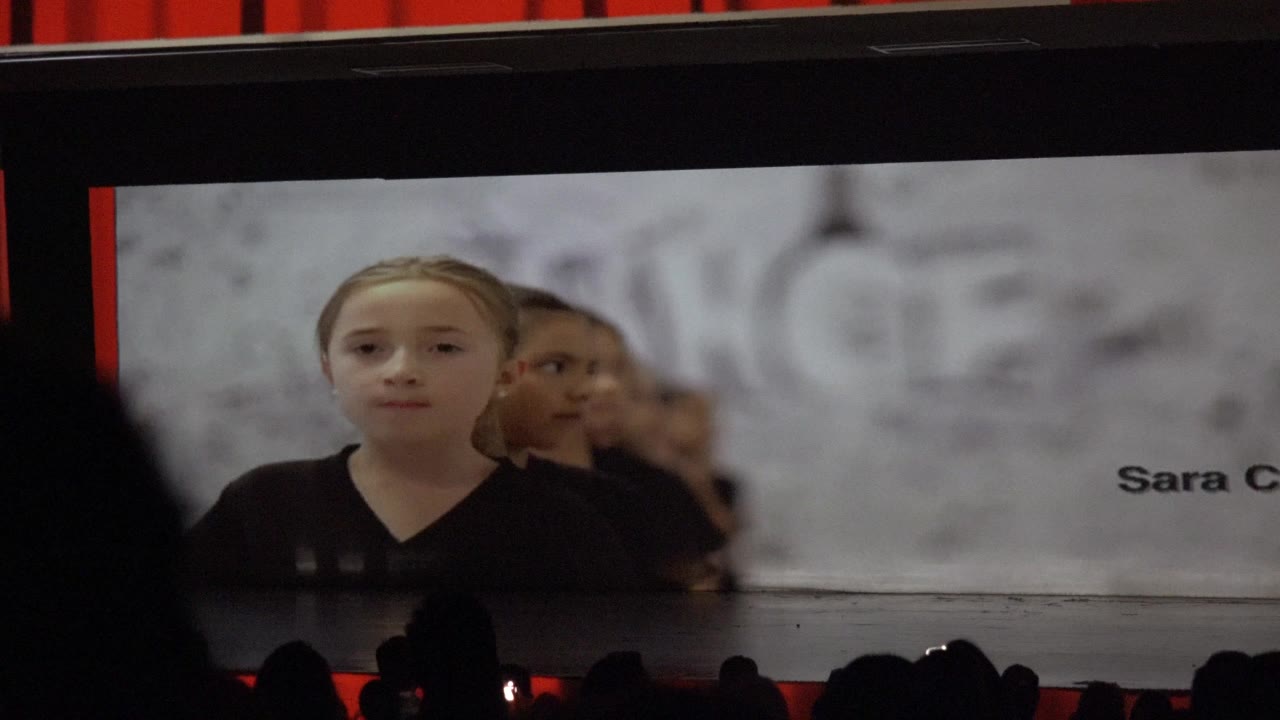 A young child appears on a large stage screen in San Jose Del Cabo BCS Mexico, with dark audience silhouettes low in frame and red curtains above.