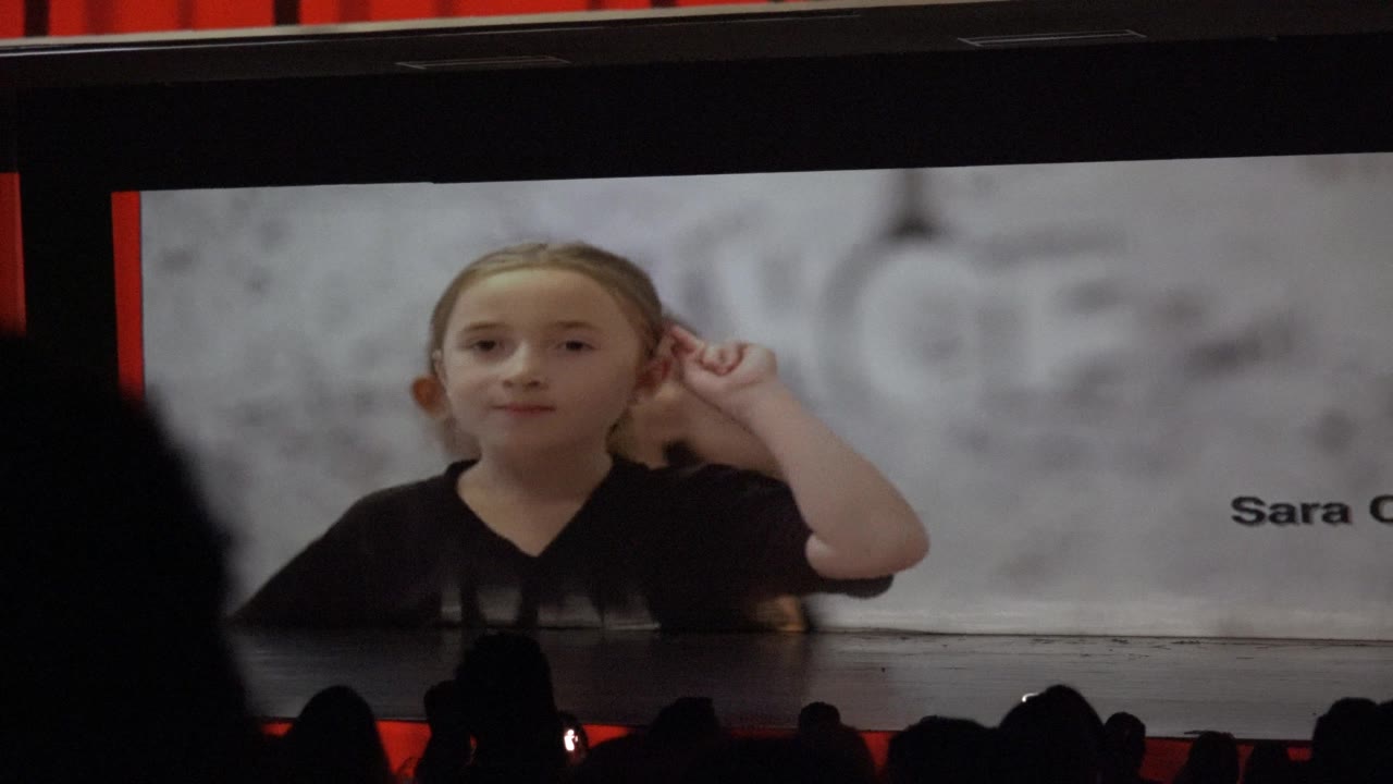 Child raises a hand near her ear on the stage screen.