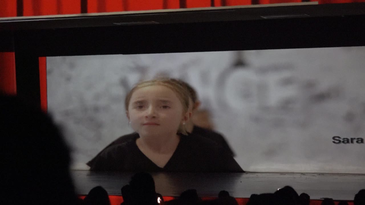 A child leans slightly forward on the large screen as the name text sits near the right edge, with audience heads silhouetted below in San Jose Del Cabo BCS Mexico.