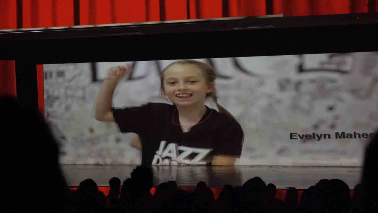 A projected young girl in a black dance shirt appears on a large screen in San Jose del Cabo BCS Mexico while dark audience silhouettes sit in the foreground.