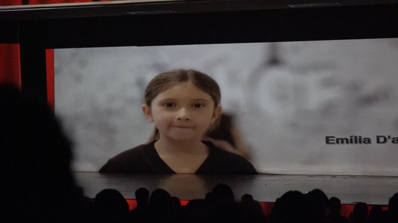 In San Jose Del Cabo BCS Mexico, a girl remains on the projection screen as partial name text appears to the right and audience silhouettes remain below.