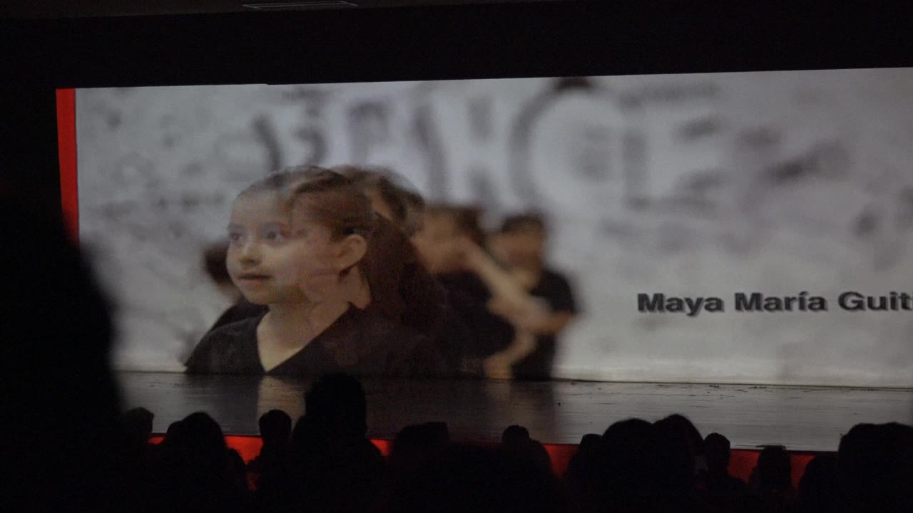 In San Jose del Cabo, Mexico, a child in a black shirt appears on a stage screen beside the name text Maya Maria Guit, with audience silhouettes across the bottom of the frame.