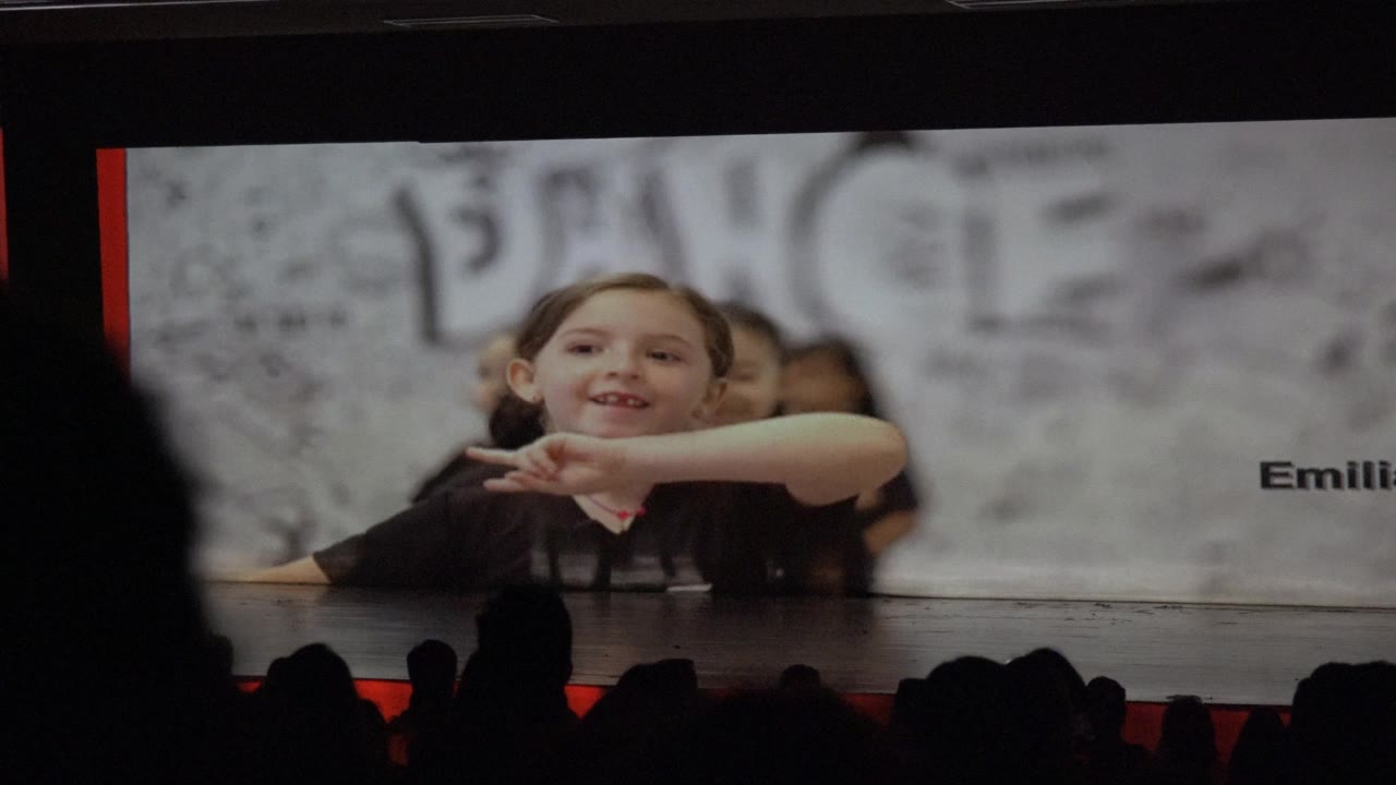 Smiling child gestures on stage screen above audience silhouettes