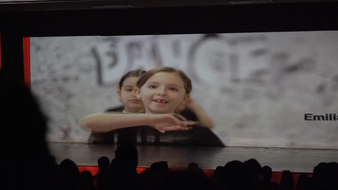In San Jose del Cabo, Mexico, a child faces forward on the stage screen with another child partly visible behind, name text cropped at the right, and audience silhouettes below.