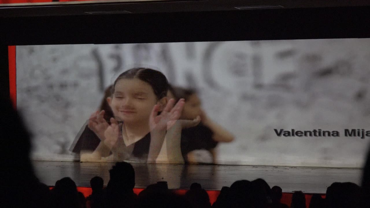 In San Jose Del Cabo BCS Mexico, a child in a black outfit waves near a projected name graphic while audience silhouettes line the bottom of the stage view.