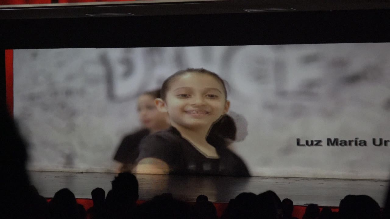 In San Jose Del Cabo BCS Mexico, a child in a black outfit smiles near the right side of the frame, with another performer partly visible behind her and projected name text cut off at the edge.