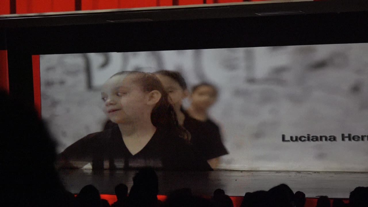 In San Jose Del Cabo BCS Mexico, a child in a black shirt appears on a large stage projection while audience heads form dark silhouettes along the bottom of the frame.