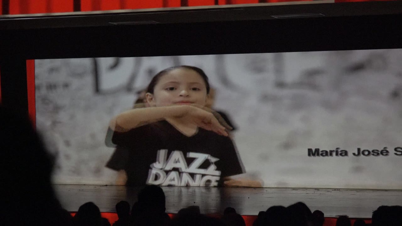 A child dancer in a black jazz dance shirt raises an arm across her chest on the projected screen, with the stage curtain above and audience silhouettes below in San Jose Del Cabo BCS Mexico.