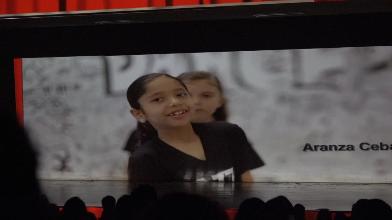 Two children in black dance shirts face the camera on a stage in San Jose del Cabo, Mexico, with dark audience silhouettes across the lower foreground.