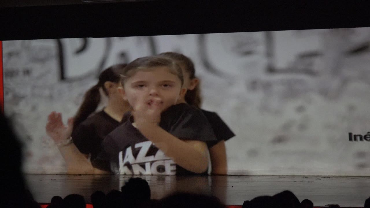 A young dancer gestures near her face while another child stands behind her, with the gray dance backdrop and audience silhouettes visible in San Jose del Cabo, Mexico.