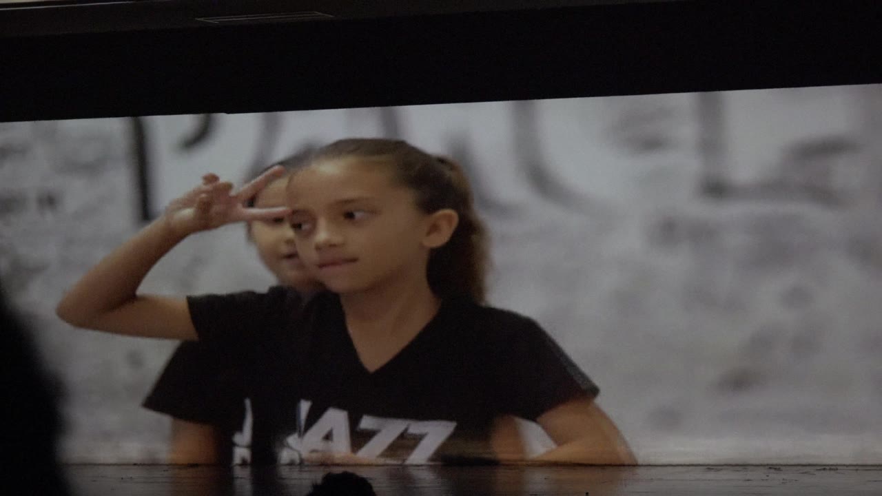 A child in a black shirt makes a peace sign behind a glossy table in San Jose del Cabo, Mexico, with another child partly visible behind her and a gray marked wall in the background.