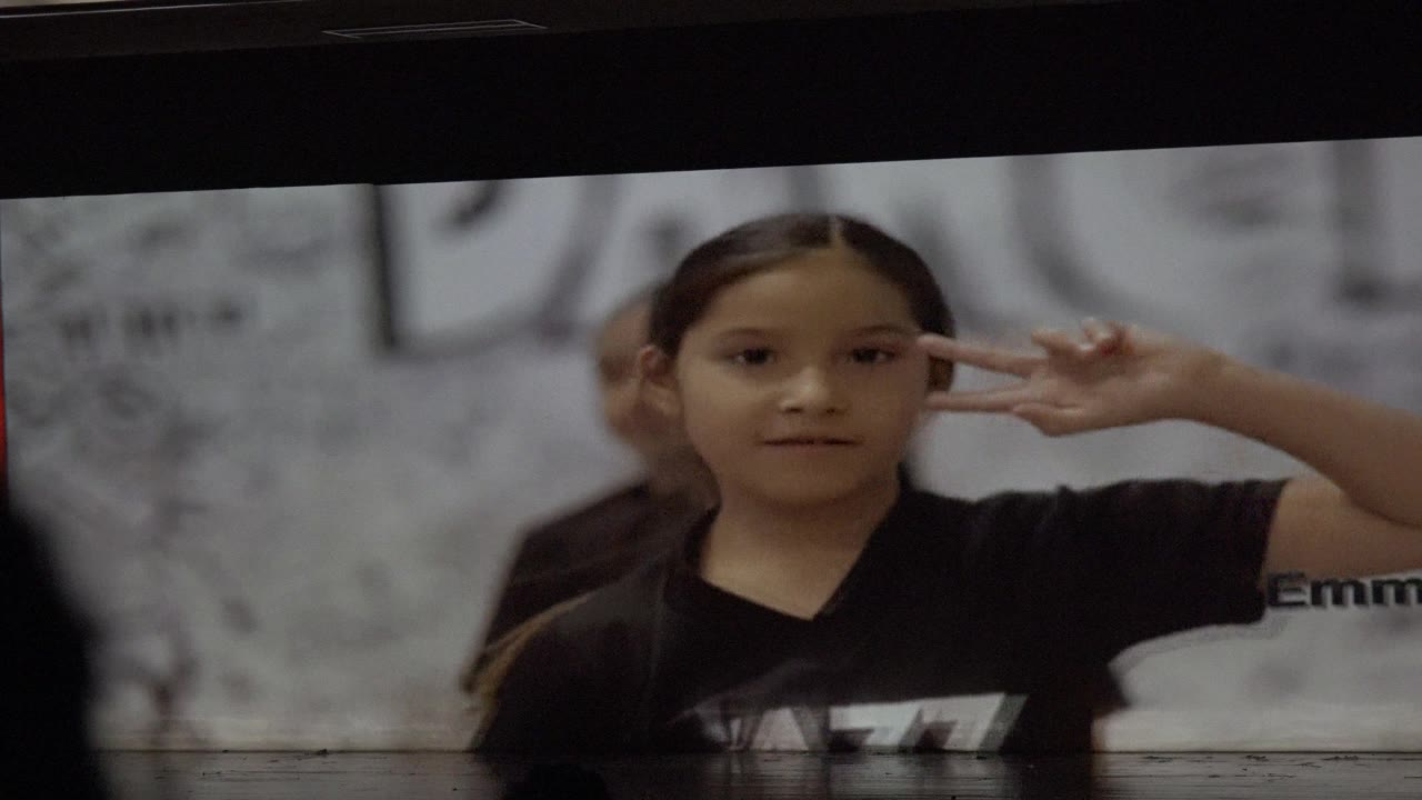 A child in a black shirt raises a peace sign near her face in San Jose del Cabo, Mexico, with another child partially visible behind her and gray wall text off to the side.