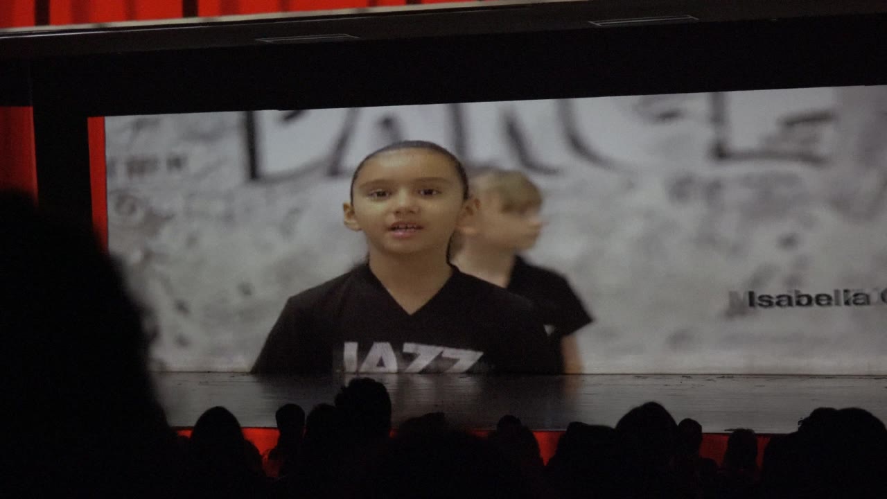 Child in black shirt centered on a large screen above audience silhouettes.