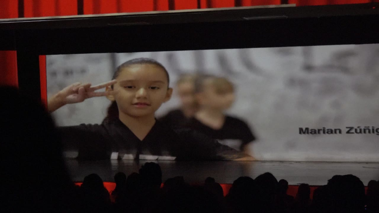 A child raises two fingers near the temple on the large screen, with another child blurred behind and audience silhouettes crossing the lower frame.