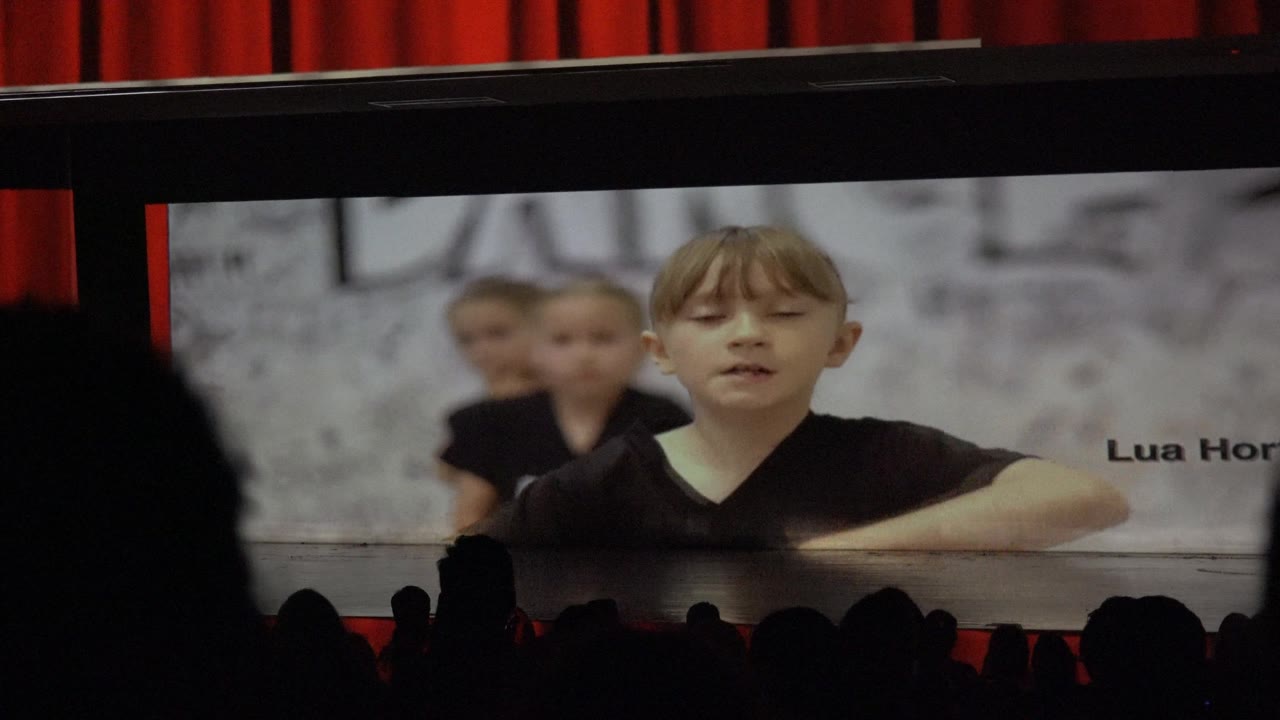 A child dancer appears on a projected stage image in San Jose Del Cabo BCS Mexico, with another child behind, red curtains above, and audience silhouettes across the lower frame.