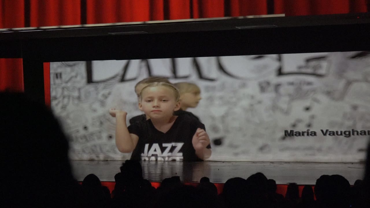 A young dancer in a black jazz dance shirt faces the audience in San Jose Del Cabo BCS Mexico, with another child behind, a dance wall graphic, red curtains, and silhouetted viewers.