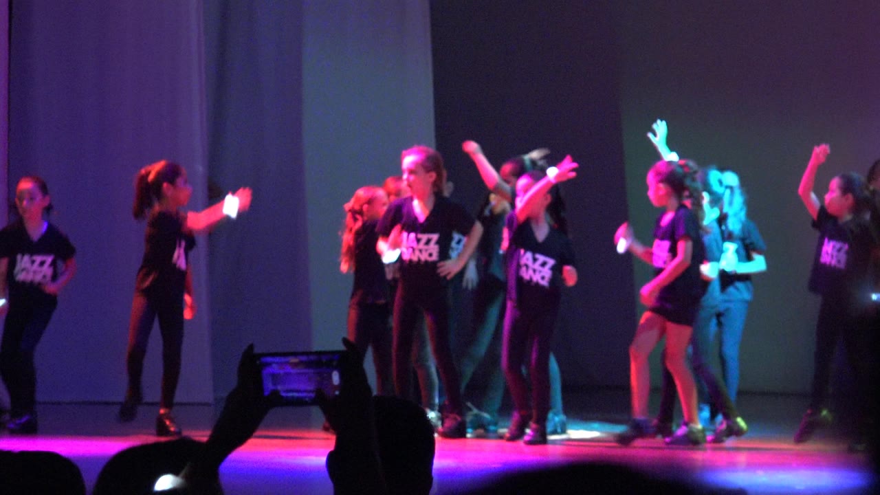 Young jazz dance group performs on a stage in San Jose Del Cabo BCS Mexico, with purple and pink lighting, black shirts, and audience phones silhouetted at the bottom.